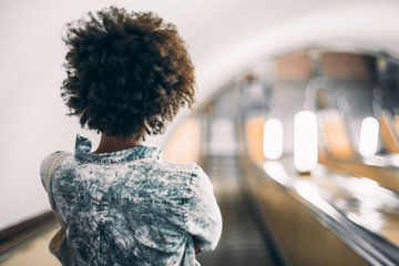 Rear view of black teenage girl with curly afro hair in trendy blue shirt standing on descending escalator in subway with copy space for young advertising text message or promotional content