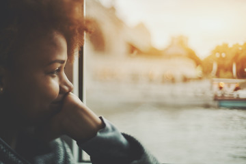 True tilt shift view of smiling black teenager girl near window traveling by ship, biracial young female is laughing and covering her mouth with hand while sitting inside of small cafe near water - Powered by Adobe