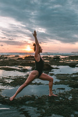 Fototapeta na wymiar Attractive red hair woman practicing yoga on the coast of Indian ocean in a black swimsuit during sunset, Indonesia, Bali