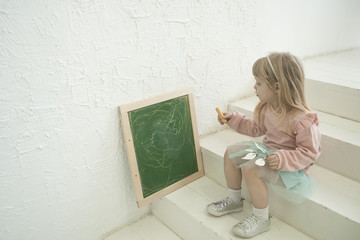 Cute toddler girl in silver head bound writing on chalk blackboard, sitting