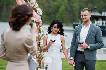 Young fashionable lovely couple with speech master of wedding ceremony against decor arch on wedding anniversary and marriage proposal. Vine glasses at hands.