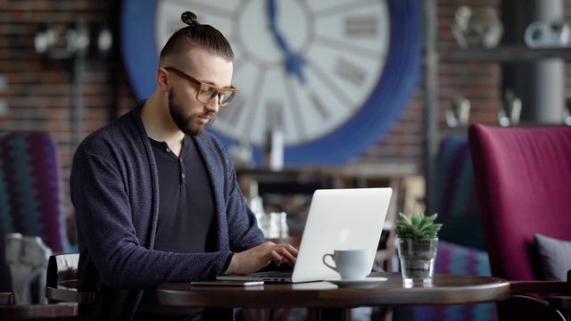 A serious writer prints on the keyboard of the laptop the text of the documentary novel that came to mind, a man with bad eyesight in glasses is sitting in a restaurant with a cup of coffee