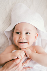 beautiful smiling newborn baby boy covered with white bamboo towel with fun ears. lies on a white knit, wool plaid bright interior. The mom's hand is on the child. The natural light