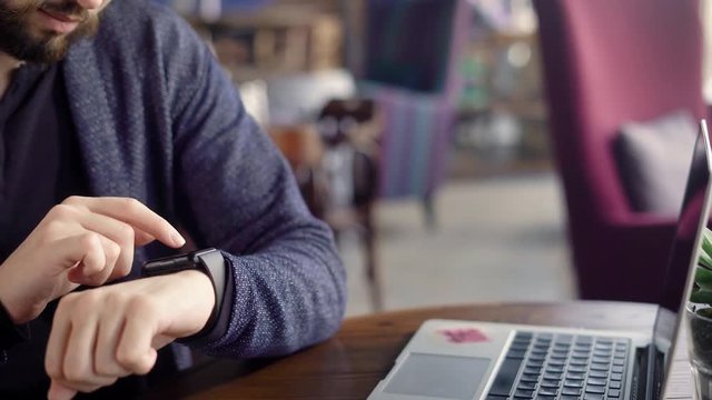 A young hipster with glasses to create a style uses smart watch in order to check the mail of social networks, on a wooden table in a restaurant is a laptop