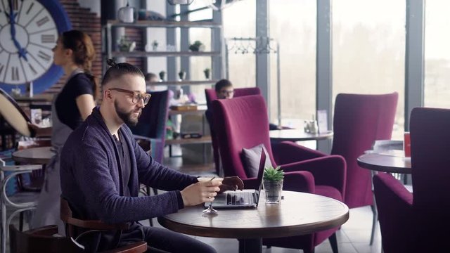 A woman waiter brought a cocktail on a tray for a stylish hipster, who has hair removed in a ponytail, a man browses fashion blogs on a laptop in a trendy restaurant