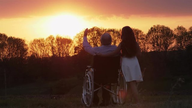 Granddaughter with invalid grandfather in a wheelchair at sunset. The man enjoys the sunset.