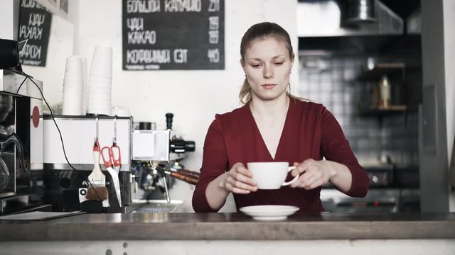 Portrait of a young woman barista putting a coffee cup and a saucer on a wooden counter and smiling. Locked down slow motion medium shot