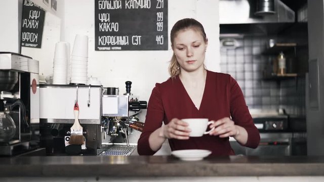 Portrait of a young woman barista putting a saucer and a coffee cup on a wooden counter and smiling. Locked down slow motion medium shot