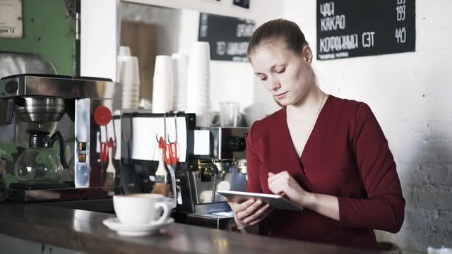 Portrait of a woman barista using a tablet computer standing behind the counter in a coffee shop. Locked down real time medium shot