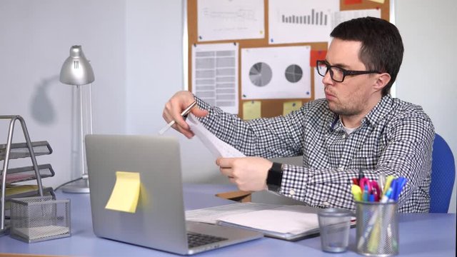 The head of a small firm checks the documents with a serious look, emphasizing the important points. A man is sitting in the office at the desk with a laptop.