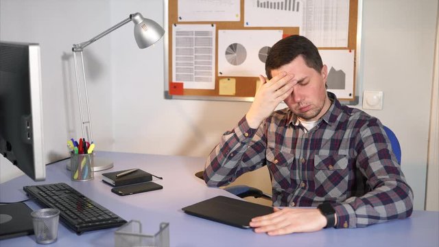 The man who sits behind the workplace holds his hands to his head, he has a headache. A man worked hard in the office, using a mobile phone, tablet, computer, now he needs a break and rest