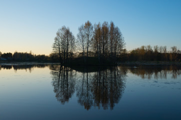 Fototapeta na wymiar Calm sunset on the lake with island covered by group of trees and horizon in the middle of frame