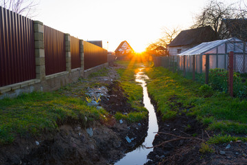 Small ditch between fence in the village leads to the sunset sun