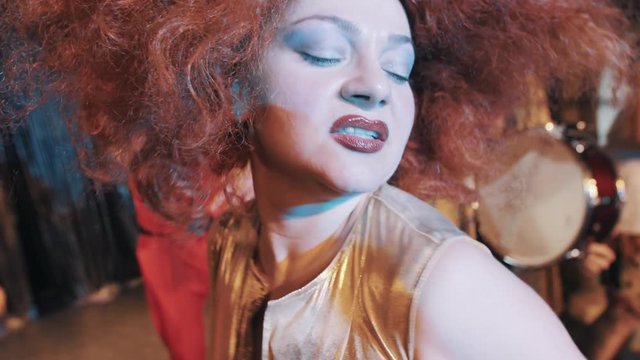 Woman in puffy carroty hair wig grimacing for camera and dancing on scene with troupe, close up