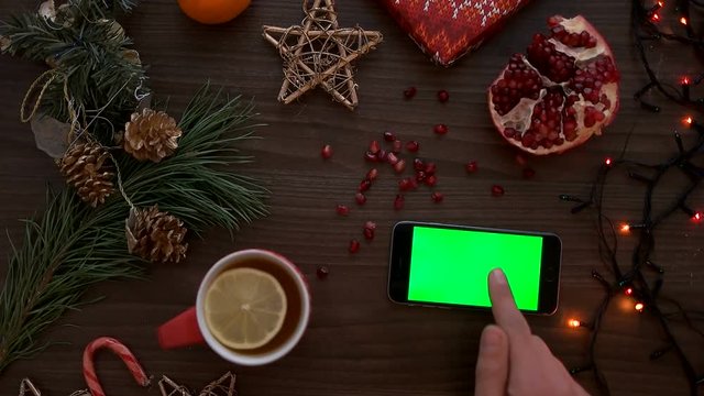 Christmas top view. Man hand using smart phone with green screen. Finger scrolling pages down on touchscreen. Beautiful christmas details on wooden table background. Chroma key. View from above