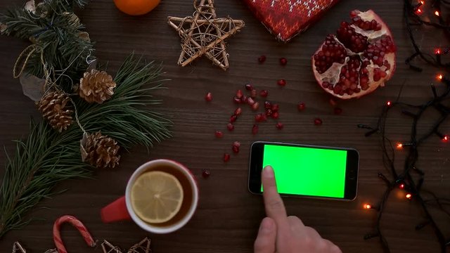 Top view man hand using mobile phone with green screen. Finger scrolling pages on touchscreen. Christmas details on wooden table background. Chroma key. Horizontal smart phone