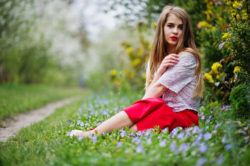 Obraz premium Portrait of sitiing beautiful girl with red lips at spring blossom garden on grass with flowers, wear on red dress and white blouse.
