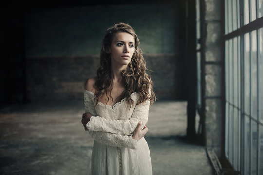 Beautiful girl in in white vintage dress with curly hair posing near the loft window. Woman in retro dress. Worried sensual emotion . Retro fashion