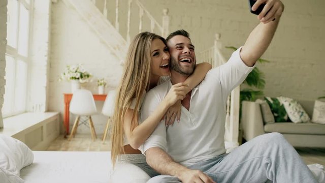 Young beautiful and loving couple take selfie picture on smartphone camera and kiss while sitting in bed at the morning