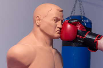 Red boxing gloves punching a dummy mannequin doll on a gym with a blue punching bag on the background