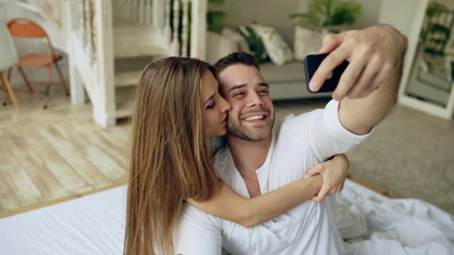 Closeup of young beautiful and loving couple take selfie picture on smartphone camera and kiss while sitting in bed at the morning