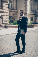 Full size photo of an intelligent stylish bearded man in a suit and glasses outdoors. He is holding his tablet, fixing a tie