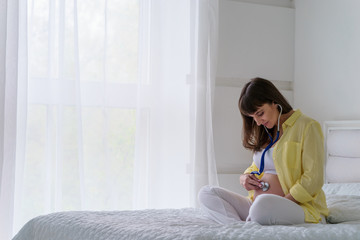One european pregnant woman with smile is listening heart beat of unborn child inside her tummy by stethoscope while she sitting on white bed in light bedroom at sunny day