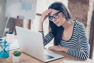 Tired overwokred young businesswoman in glasses sitting at the table and touching her forehead