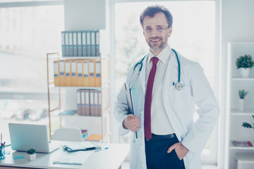 Happy successful doctor in glasses standing at the office, holding clip board. His workplace is bright and modern