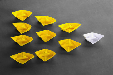 Leadership concept white leader paper boat standing out from the crowd of yellow boats on black wood backgroud