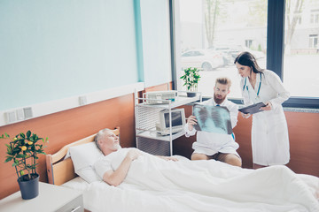 Young red bearded doc is showing x ray to the nurse and telling about the surgery, patient is aged, he is in the bed upset