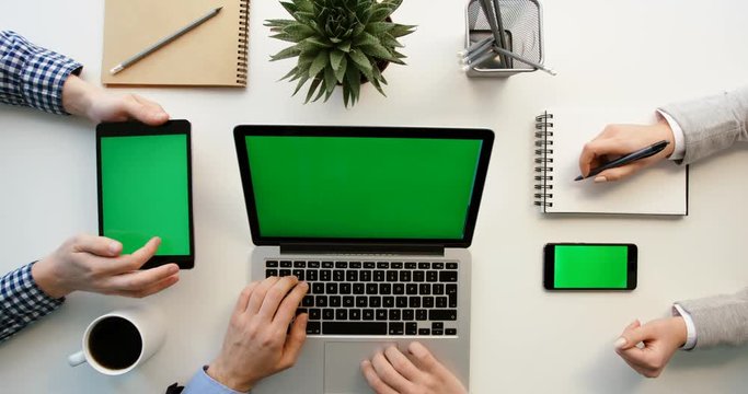 Business team working together at office desk and checking a financial report, men using tablet and laptop with green screen and woman using smartphone and writing in notebook. Hands top view. Green