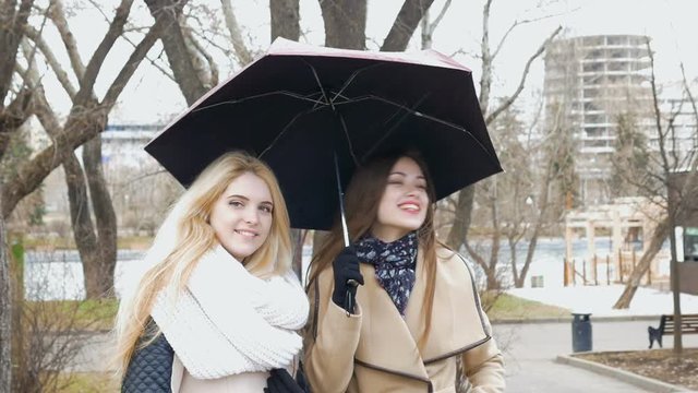 Two beautiful girlfriends blonde and brunette - talking standing under an umbrella. Sunny day, the wind blows.