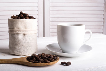 Cup of coffee, coffee beans in the bag, wooden spoon on a wooden white background table. Side view. Place for text or inscription.