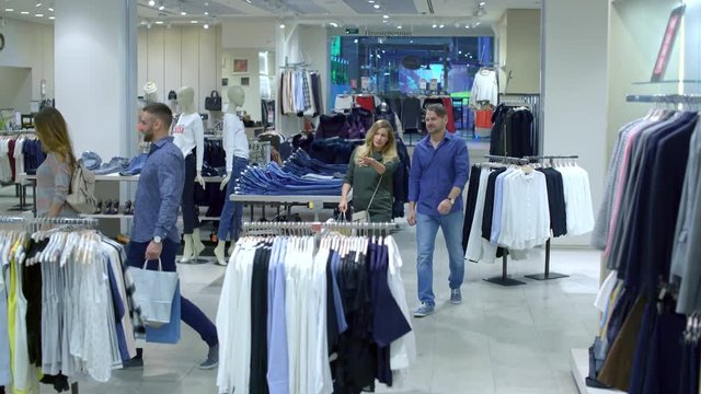 Wide shot with tilt up of clothing store: young women and men walking excitedly between clothes racks and looking at new collection 