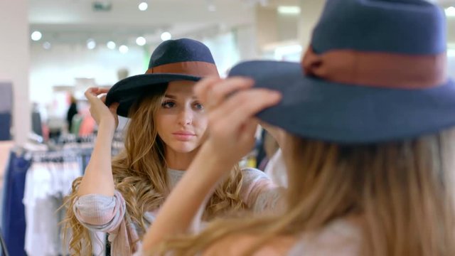 Medium shot of beautiful blond woman looking in mirror in clothing store and trying on hat