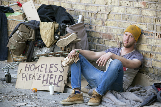 
Sad homeless man with  cardboard, sits near  wall. Carriage is home
