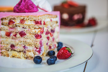 Ассорти из больших кусков разных тортов: три шоколада, морковный, клубничный, шоколадный. Торты украшены ягодами, зефиром, покрыты разноцветным кремом. Кусочки тортов стоят на тарелках на белом столе.