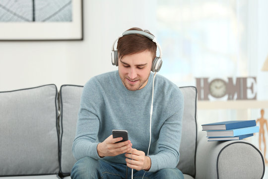 Concept of audiobook. Handsome young man with headphones and phone sitting on sofa at home