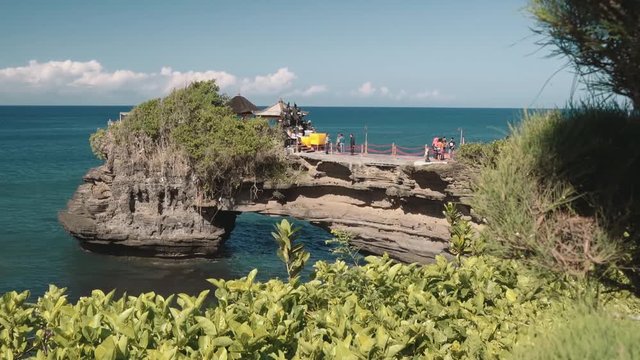 Pura Batu Bolong shrine, ( another temple in Tanah Lot area ) on headland with archway in ocean view from trees and bushes. Shot with Sony a7s on slider on sunny day with blue sky
