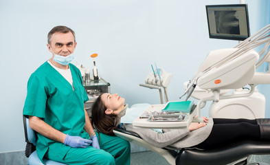 Friendly male dentist with dental equipment during treatment in the dental office. Female patient lying on the dental chair. On the background screen with X-ray the patient's teeth. Dentistry