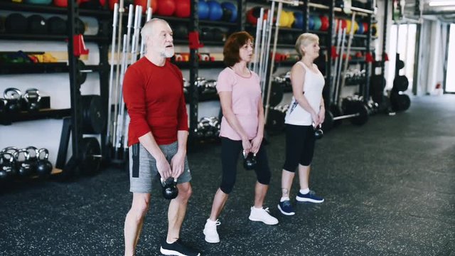Fit seniors in gym working out, doing squats with kettlebells.