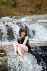 Portrait of a brunette in a black dress near a mountain river. The model is posing at the waterfall.