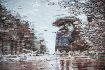 reflet dans une flaque d'eau d'un couple marchant avec un parapluie sur un place pavée