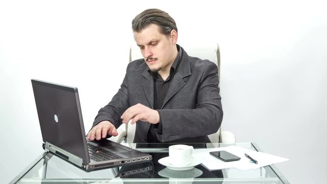 A young man is working in his office behind his computer. Then he is stopping and taking a sip of a coffee from his cup on the desk.