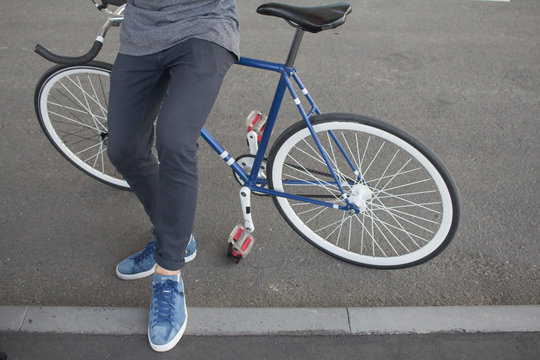 Close up picture of man sitting on his blue bicycle. Legs of man in black jeans and blue shoes sit on blue retro fixed gear bike with white wheels 