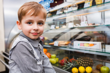 A little boy stands near a showcase with sweets and fruits in a cafe