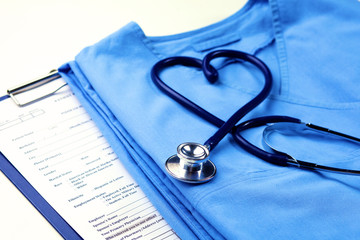 Medical stethoscope twisted in heart shape lying on patient medical history list and blue doctor uniform closeup