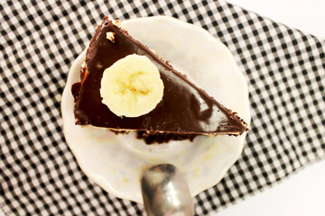A piece of chocolate cake with milk chocolate and a close-up banana lies on a porcelain plate with a metal spoon. Isolated on white background