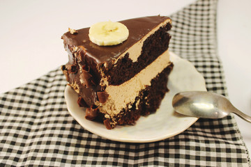 A piece of chocolate cake with milk chocolate and a close-up banana lies on a porcelain plate with a metal spoon. Isolated on white background